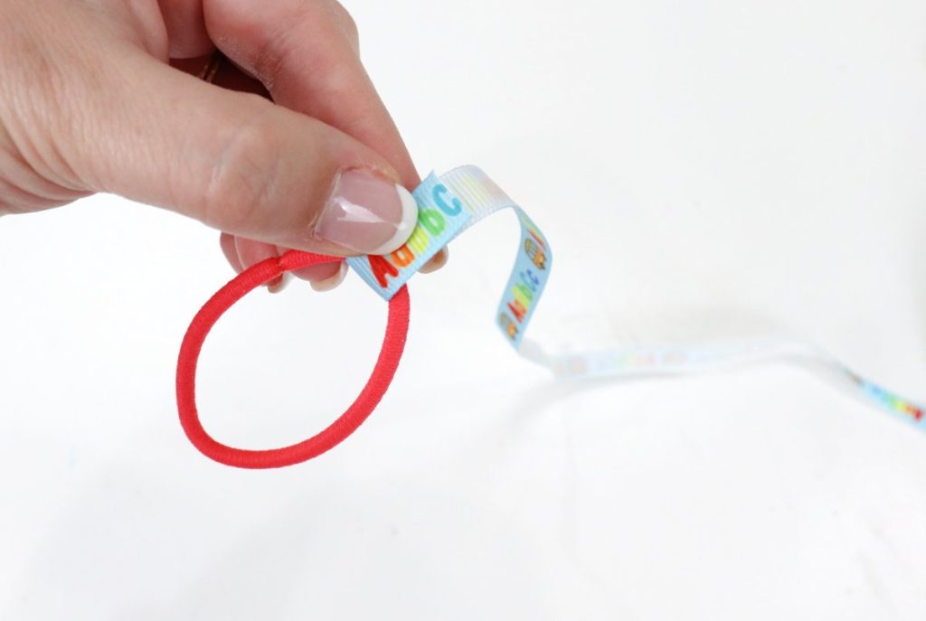 Image contains Amy’s hand holding a red hair tie with a piece of blue ribbon wrapped around it.