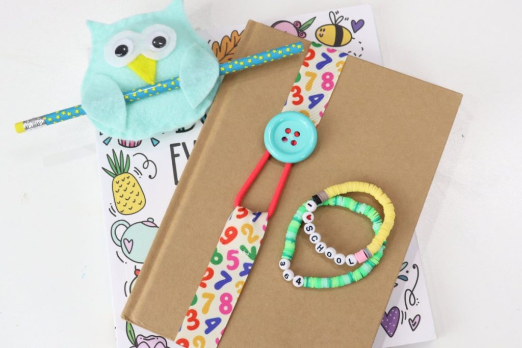Image contains a brown journal wrapped with a ribbon bookmark. Two beaded bracelets sit on top of it. The journal is on top of a copy of the book Doodle Everything, and there is a teal felt owl holding a turquoise pencil.