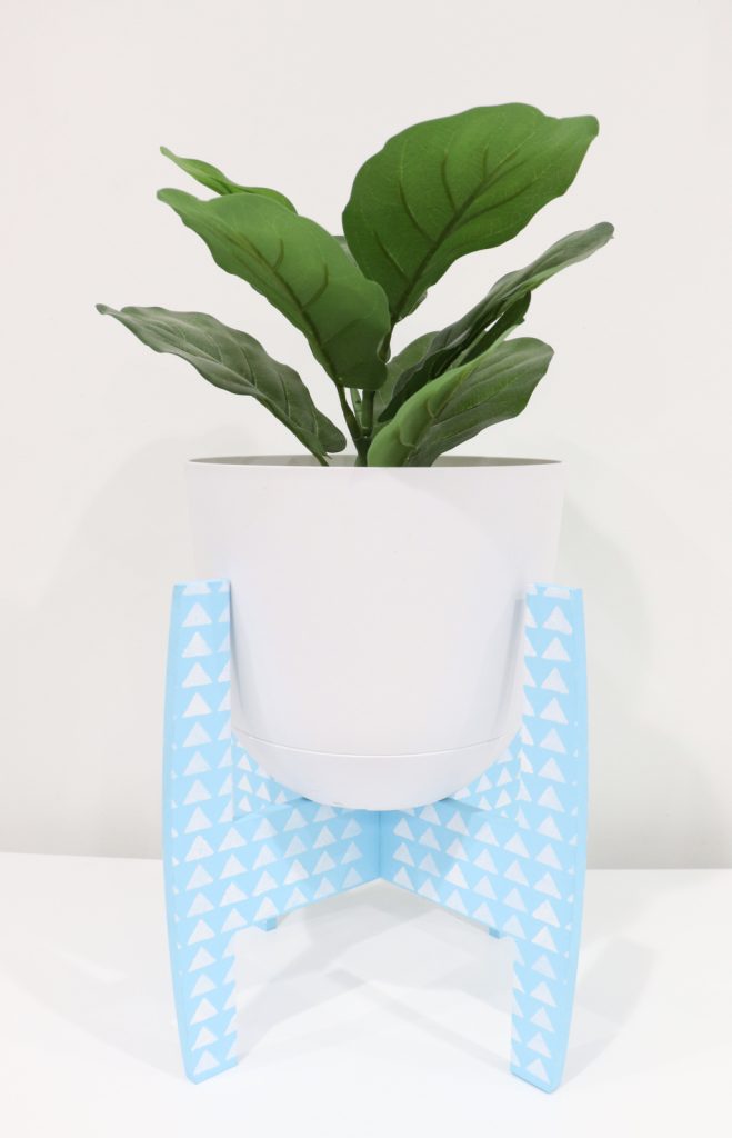 Image contains a light blue plant holder with a design of white triangles stenciled on it, holding a faux plant in a white pot.