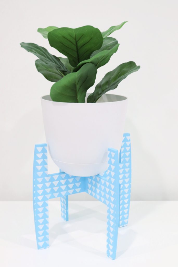 Image contains a faux plant in a white pot sitting in a blue painted plant stand with a white triangle design.
