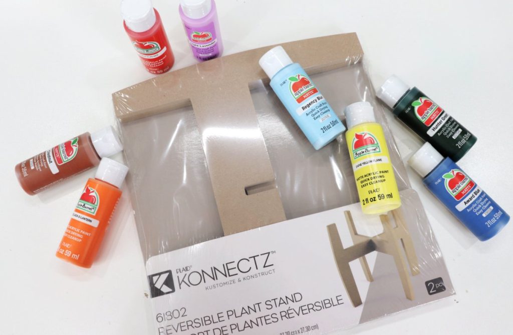 Image contains a plant stand in its packaging, and eight bottles of Apple Barrel paint in orange, brown, red, purple, blue, yellow, dark blue, and black.