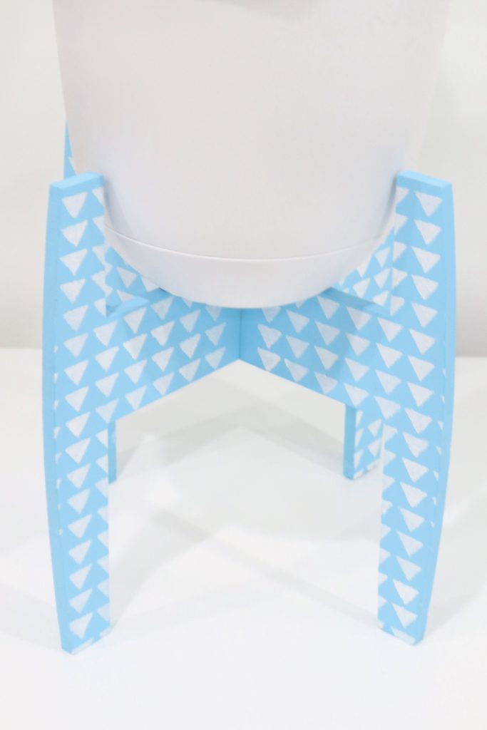 Image contains a plant stand painted light blue with a geometric triangle pattern stenciled on it in white.