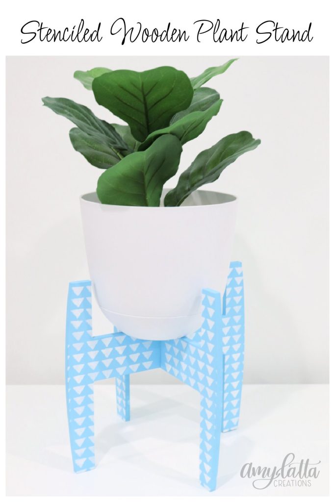Image contains a faux plant in a white pot sitting on a wooden plant stand that’s painted blue with a white stenciled triangle pattern.