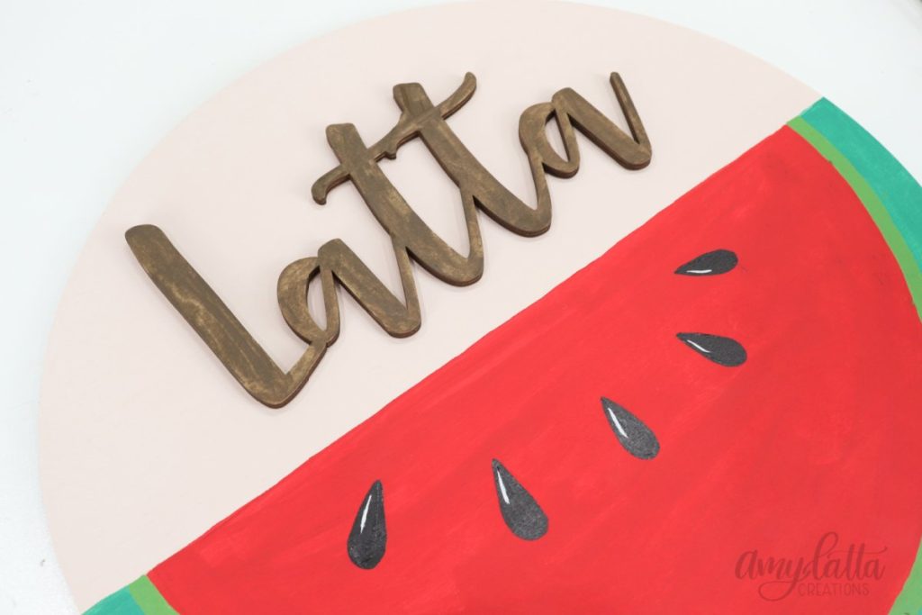 Image contains a large wooden circle. The bottom half is painted to look like a watermelon, and the top half has the name, “Latta."