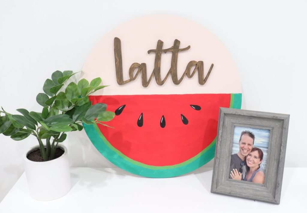 Image contains a large round wooden sign where the bottom half is painted to look like a watermelon, and the top half says the name, Latta. A faux plant and a framed photo are sitting beside the sign.