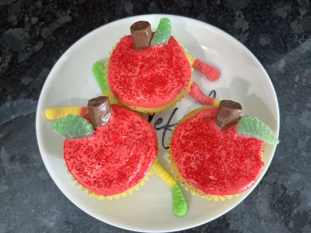 Image contains three cupcakes decorated to look like red apples on a white plate surrounded by gummy worms.