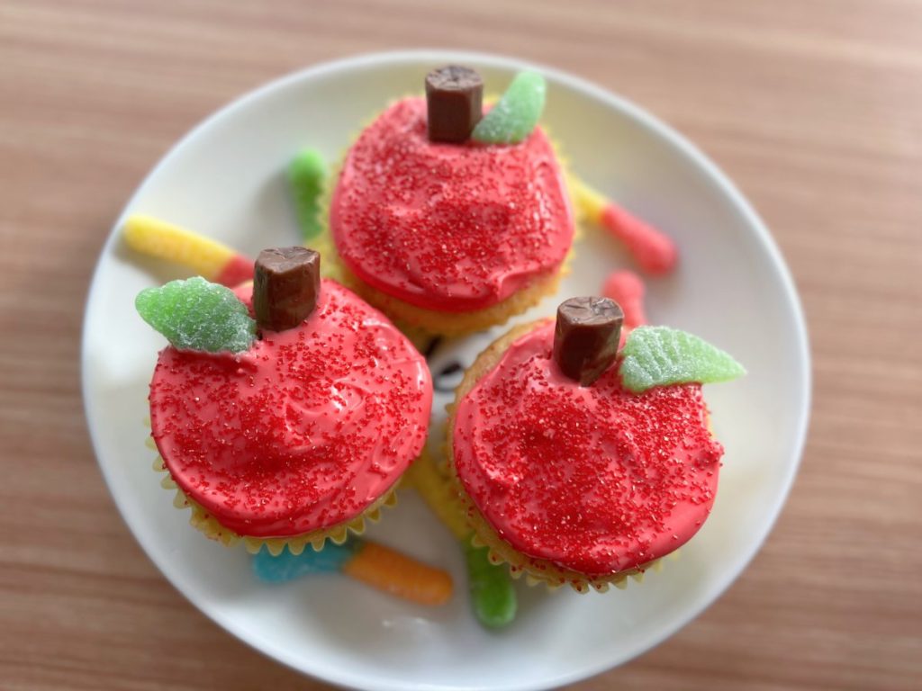Image contains three cupcakes decorated to look like red apples on a white plate, surrounded by colorful gummy worms.