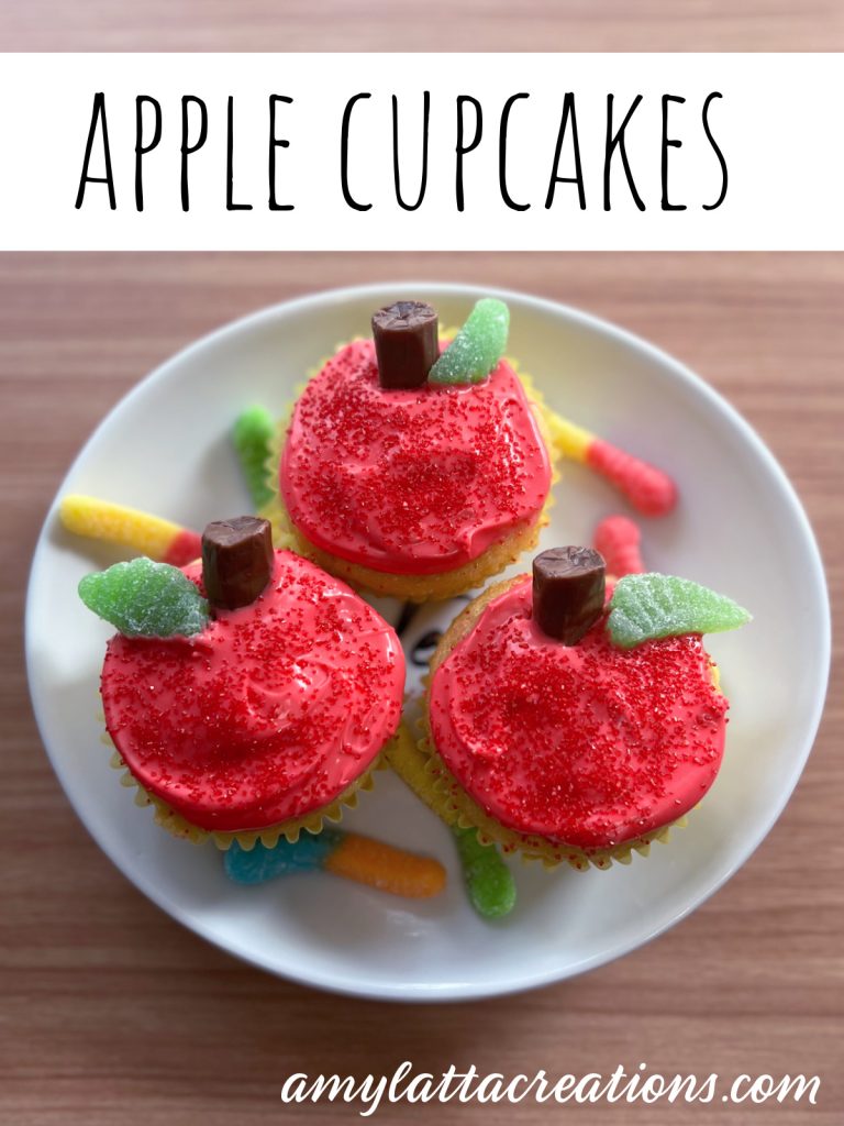 Image contains three cupcakes decorated like red apples on a white plate, surrounded by gummy worms.