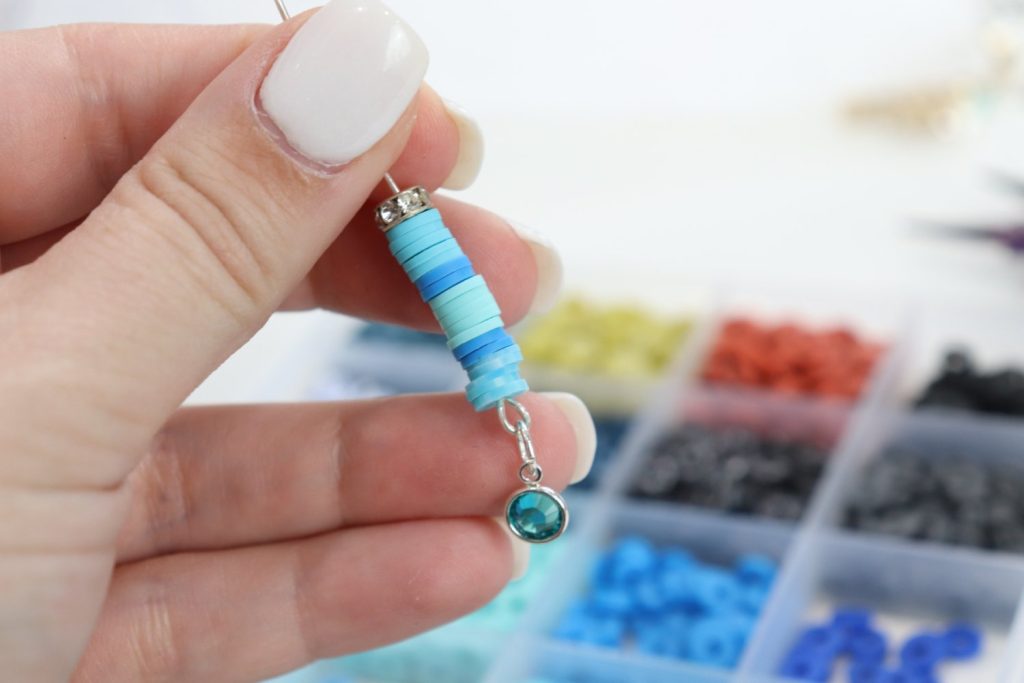 Image contains Amy’s hand holding a piece of wire with various shades of blue and teal clay beads threaded on it. A blurred tray of clay beads is in the background.