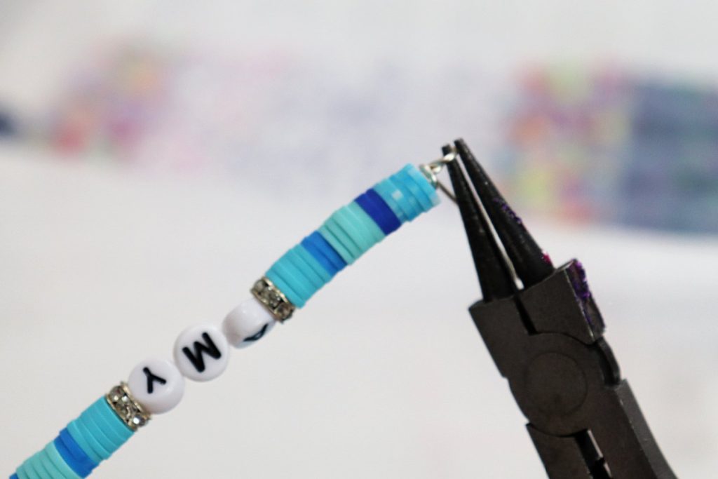 Image contains a pair of round nose pliers making a wire loop at the top of a beaded backpack charm.