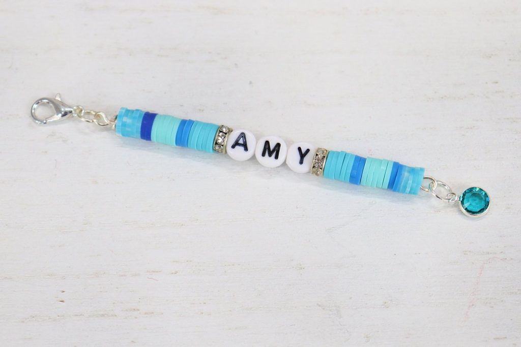 Image contains a backpack charm with the name “AMY” in the center and assorted blue and teal clay beads on either side.
