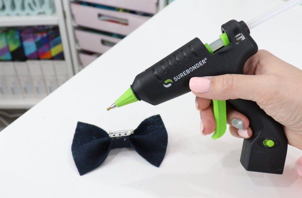 Image contains Amy’s hand holding a black and neon green hot glue gun. A felt bow sits upside down on a white table beside it.