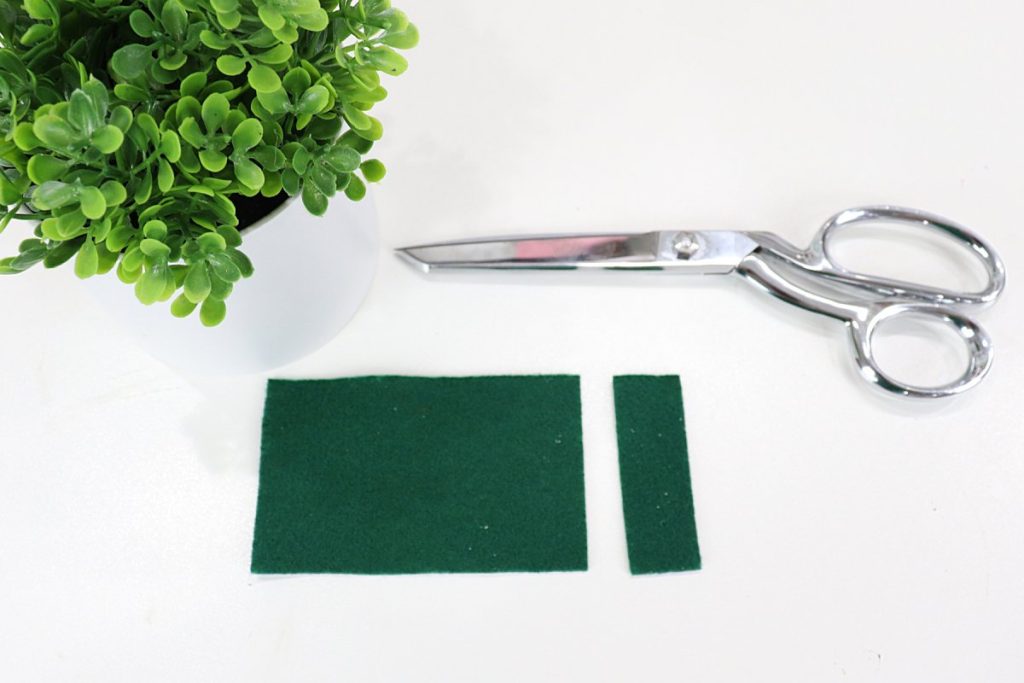 Image contains two pieces of dark green felt, a pair of silver scissors, and a faux plant on a white table.