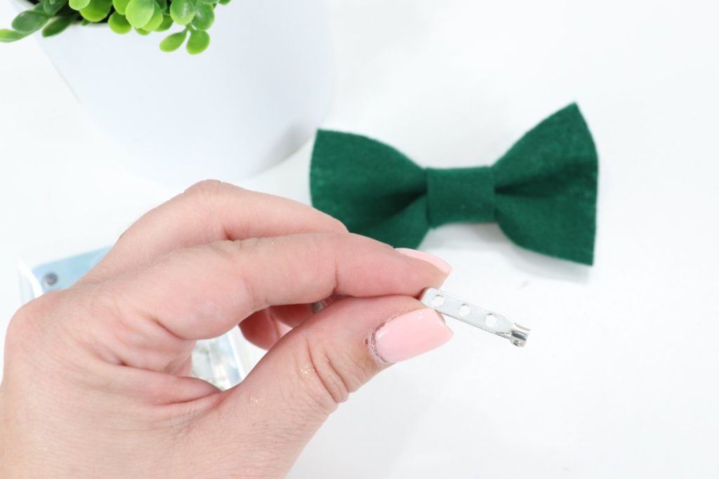 Image contains Amy’s hand holding a bar pin. In the background, a green felt bow and a faux plant sit on a white table top.
