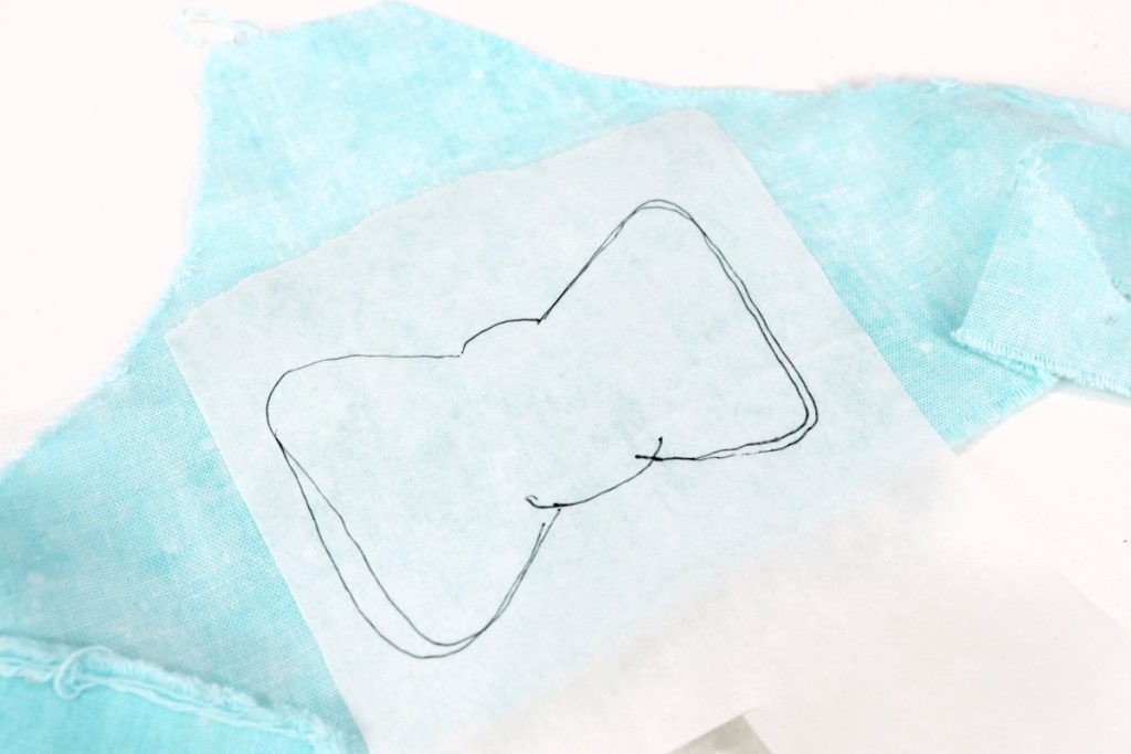 Image contains a teal fabric scrap laying face down with a piece of fusible interfacing ironed on top. A bow shape is sketched on the interfacing in black pen.