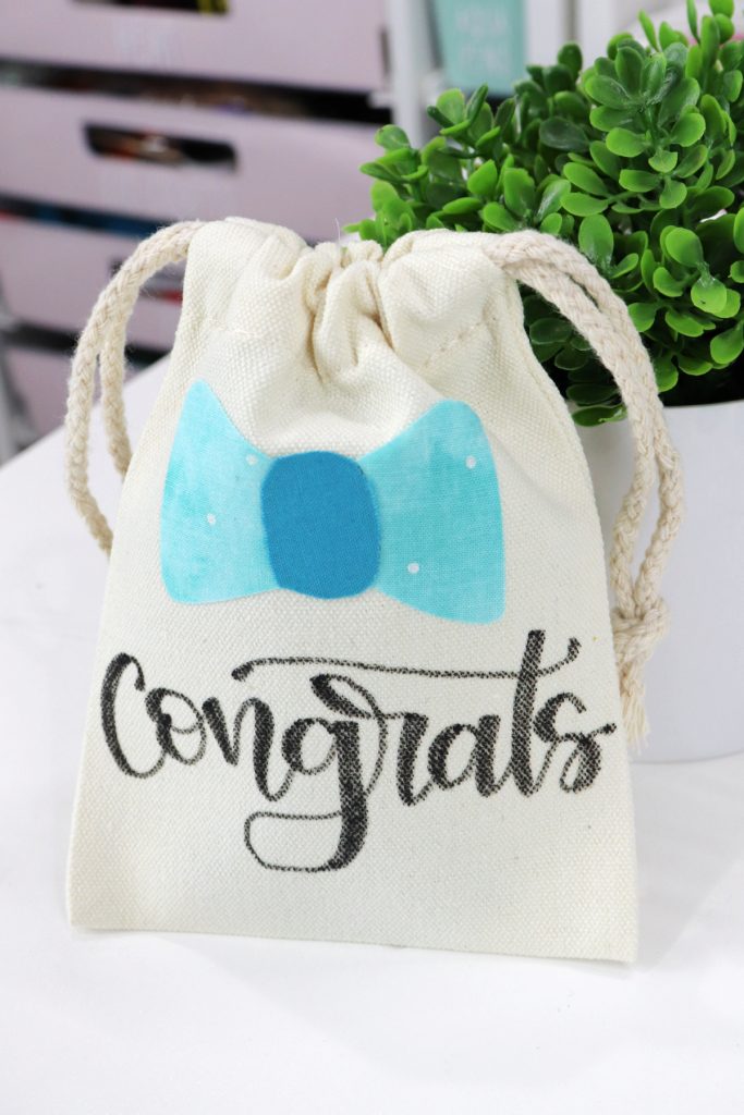 Image contains a canvas bag with a teal fabric bow and the word “congrats” lettered in black script. It leans against a faux plant on a white table.