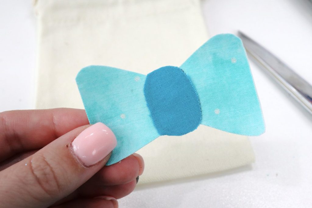 Image contains Amy’s hand holding a bow shape made of light and darker teal fabric. In the background, a pair of scissors and a canvas drawstring bag sit on a white table.