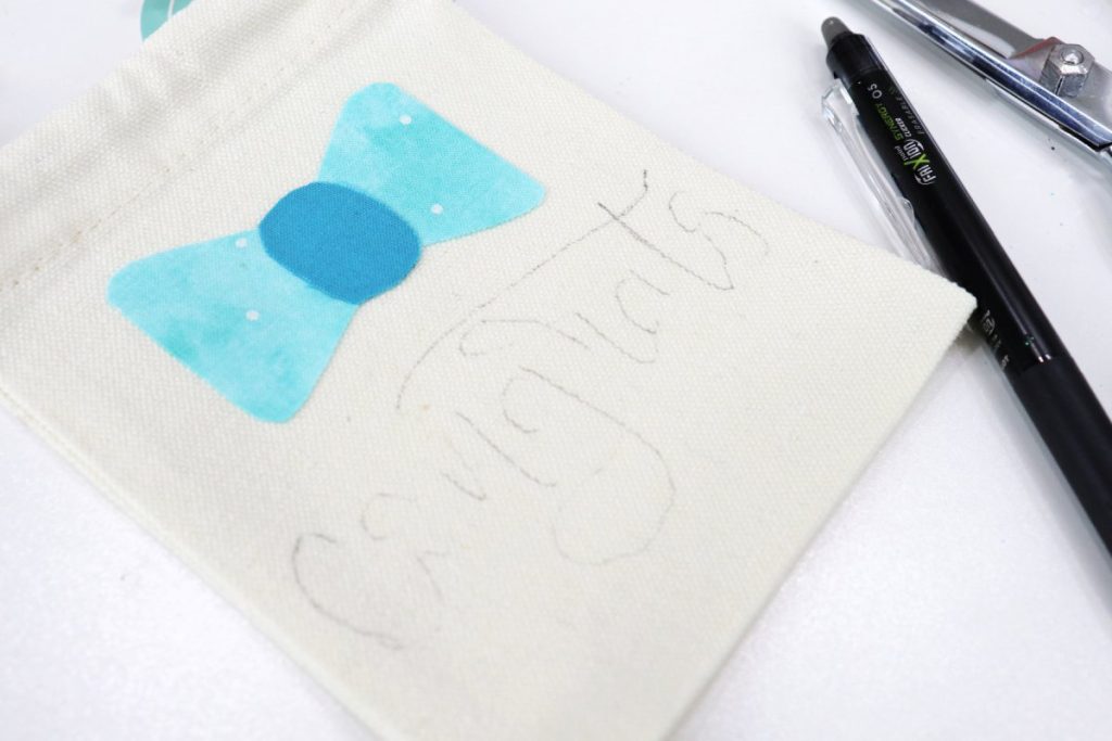 Image contains a white canvas bag with a blue fabric bow and the word “congrats” sketched in heat resistant pen. The pen and a pair of scissors sit beside it on a white table.