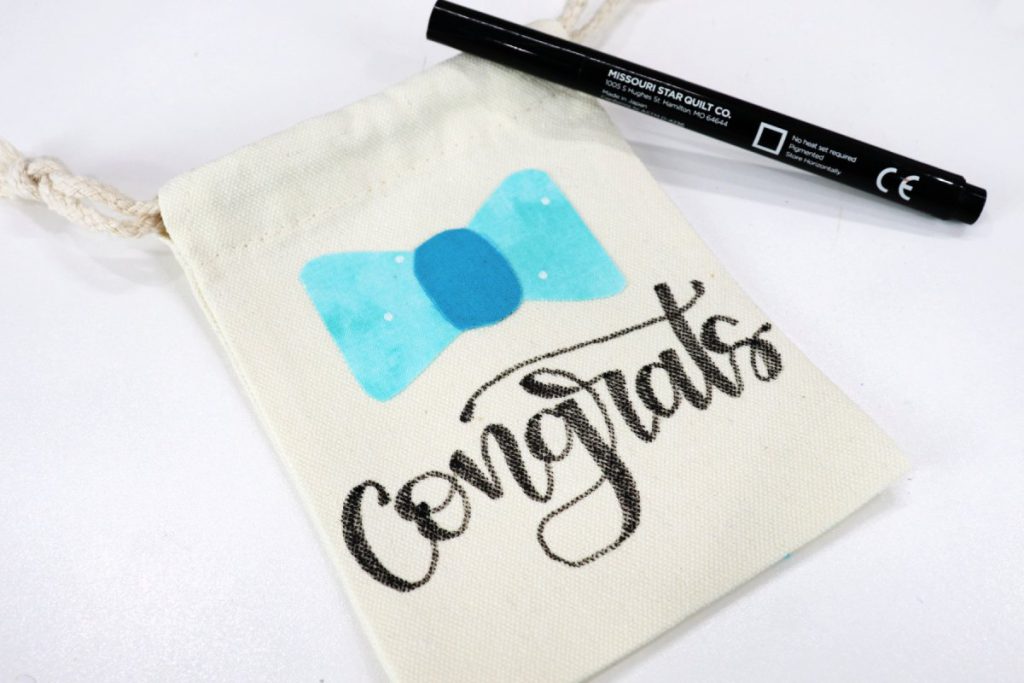 Image contains a small canvas bag with a blue fabric bow and the word “congrats’ in black script. A black fabric marker sits nearby on a white table.