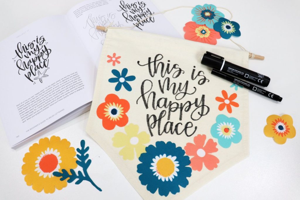 Image contains an open copy of the book “The Beginner’s Guide to Brush Lettering” with a decorated canvas banner sitting on top. A pair of black fabric markers, and assorted multi-colored iron-on flowers sit around it.