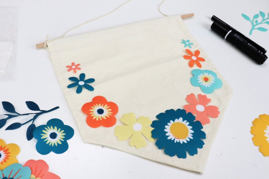Image contains a canvas banner with iron-on flowers positioned around the border. More flowers sit around it on a white background, as do two black fabric markers.