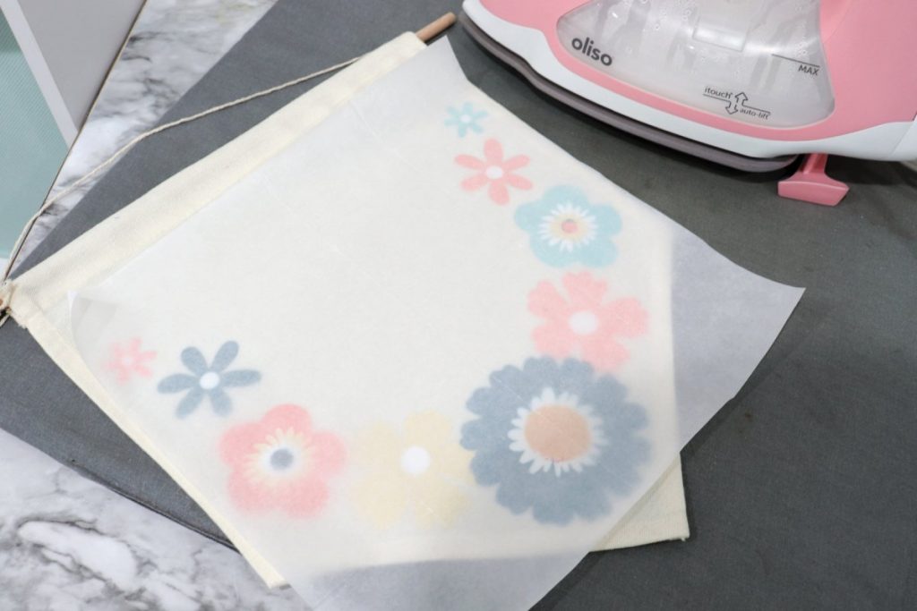 Image contains a canvas banner with iron-on flowers sitting on a grey ironing pad. It is covered with a piece of parchment paper. A pink oliso iron sits nearby.