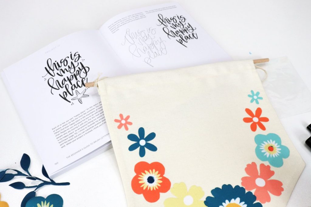 Image contains an open copy of the book “The Beginners Guide to Brush Lettering” with a canvas banner sitting on top of the pages. The banner has multi-colored flowers around the border.