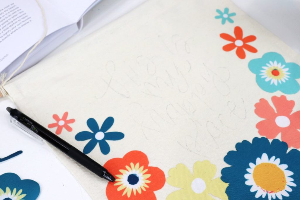 Image is a close-up of a canvas banner with multi-colored flowers around the border and a phrase sketched in the center with black pen. A heat erase pen sits on top of the banner.