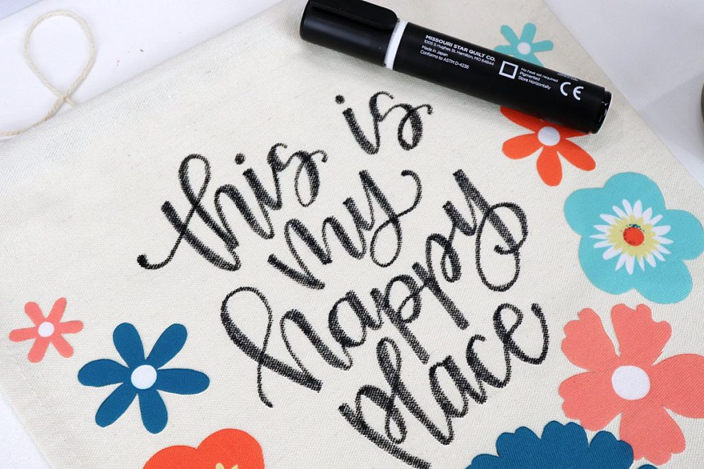 Image contains a canvas banner with the phrase “this is my happy place” hand lettered in the center with black fabric marker. Colorful flowers surround the words, and the fabric marker sits on top of the banner.