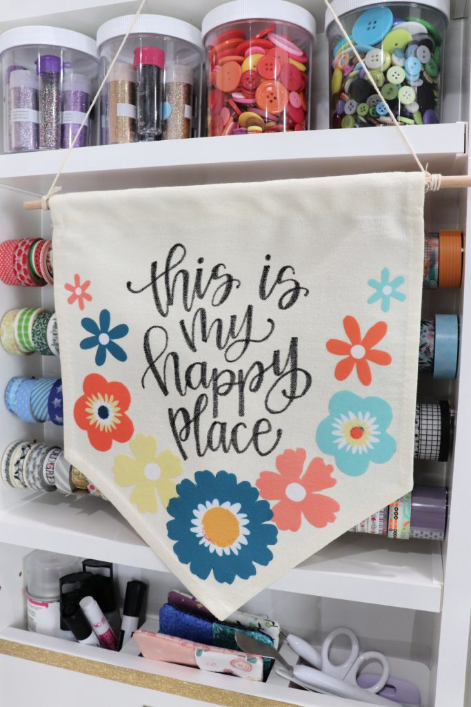 Image contains a canvas banner with iron-on flowers and the hand lettered phrase, “this is my happy place.” It hangs in front of a variety of craft supplies in jars and hanging from rods.