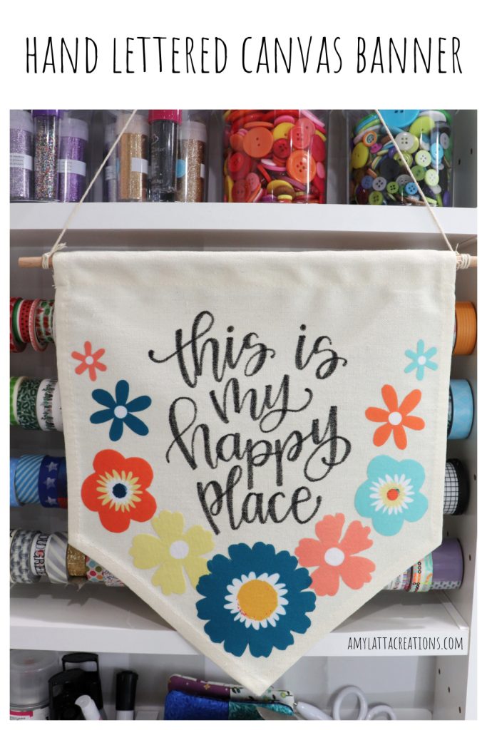 Image contains a canvas banner with multicolored flowers and the hand lettered phrase, “this is my happy place.” It hangs in front of assorted washi tape and other craft supplies.