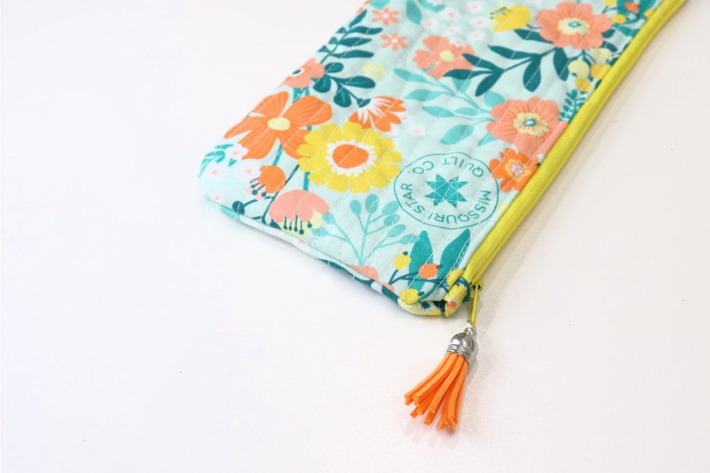Image contains a quilted teal, yellow, and orange floral pouch with an orange tassel attached to the zipper pull.