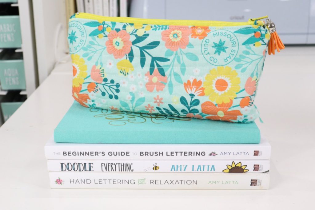 Image contains a quilted teal, yellow, and orange floral pouch with an orange tassel, sitting on top of a stack of four books on a white table. 
