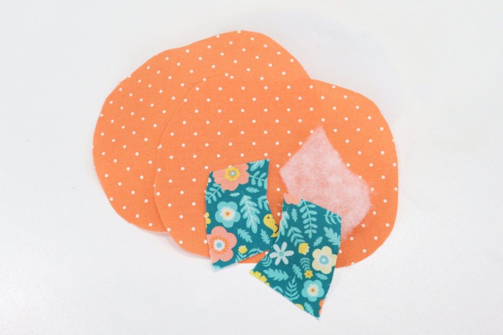 Image contains orange fabric cut into two pumpkin shapes, quilt batting, and teal fabric cut into stem shapes on a white table.