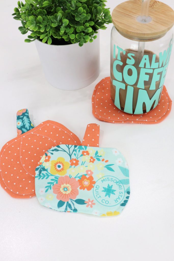Image contains a glass coffee cup with the words, “it’s always coffee time” sitting on top of an orange pumpkin coaster. To the left is a faux plant, and there are two more pumpkin coasters (one orange and one teal) in front.