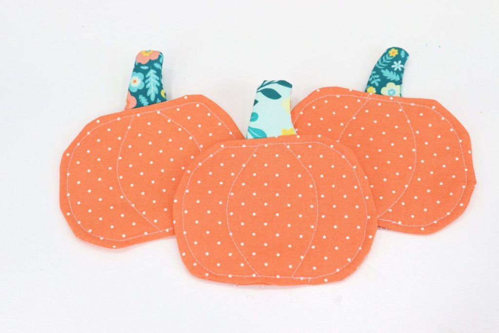 Image contains three orange fabric coasters shaped like pumpkins with stems made of teal fabric, sitting on a white table.