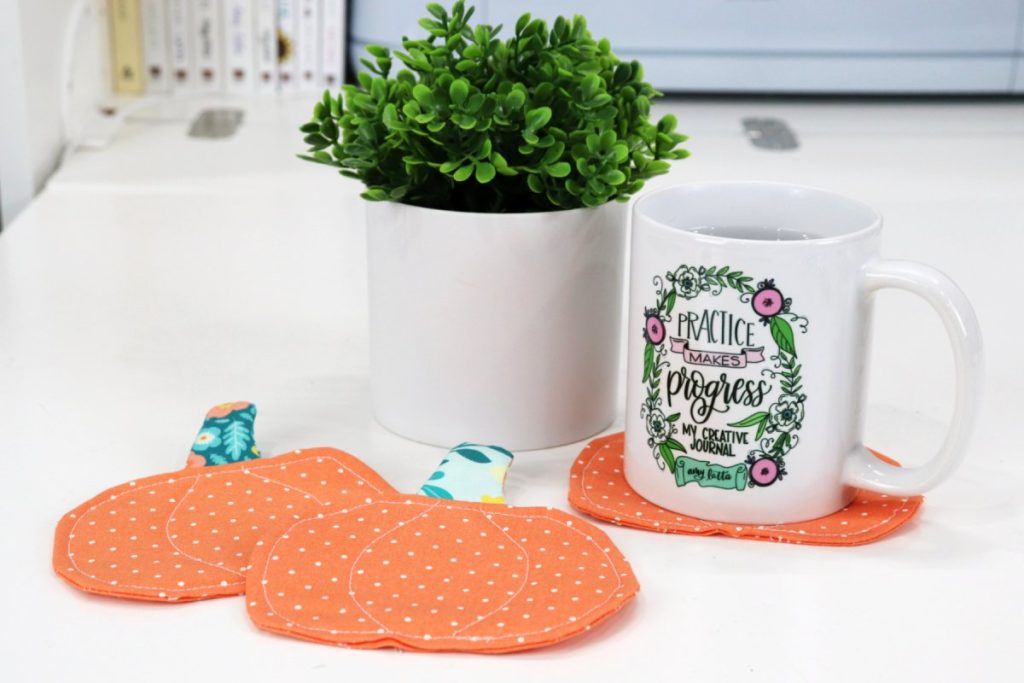 Image contains a white coffee mug sitting on an orange pumpkin coaster next to a faux plant. Two more pumpkin coasters sit in front of the mug on a white tabletop.