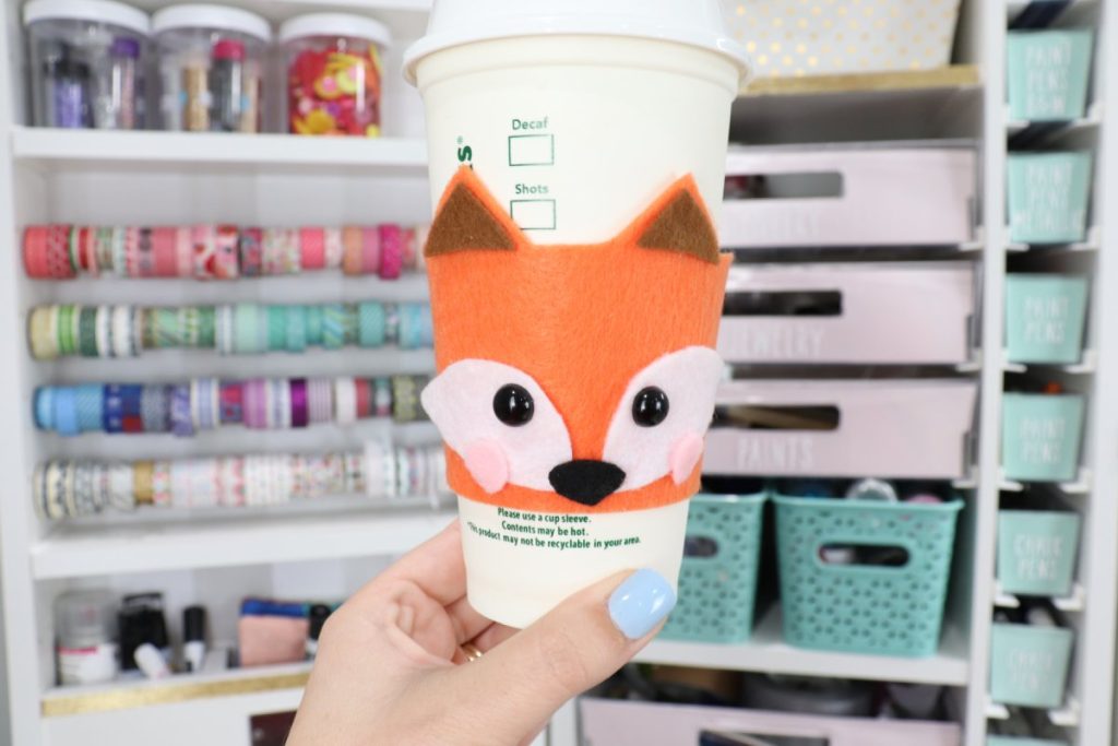 Image contains Amy’s hand holding a white to-go cup with an orange felt coffee cozy that looks like a fox.