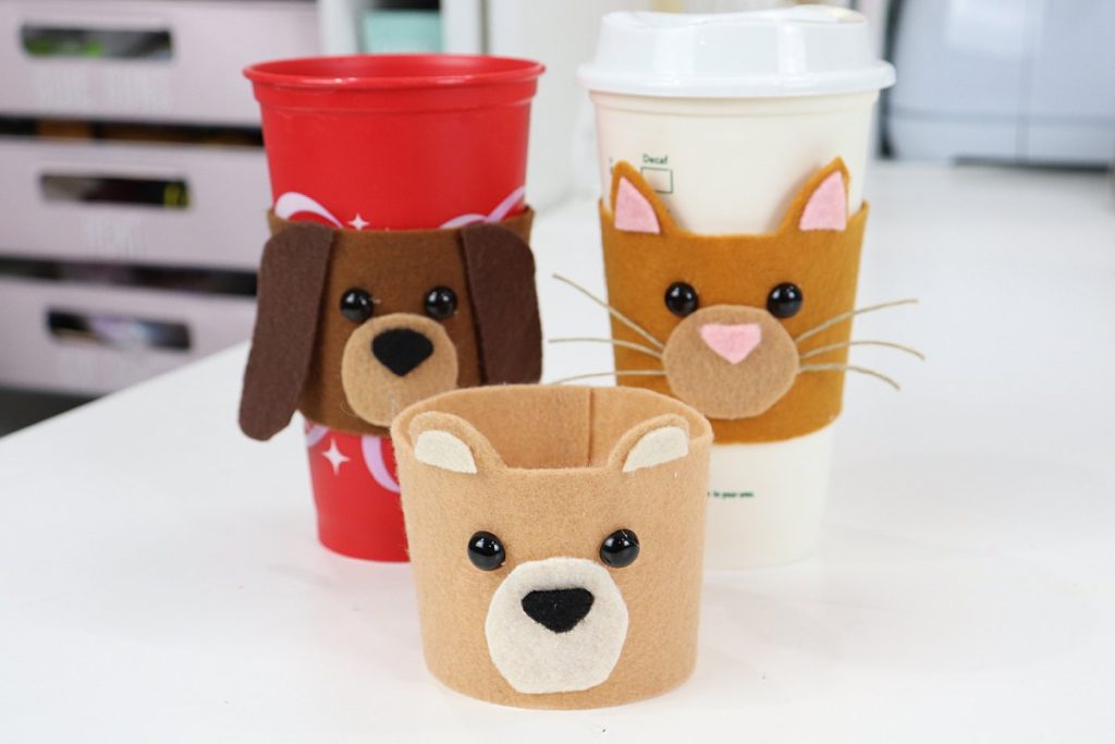 Image contains three felt coffee cozies; a dark brown dog cozy on a red cup, an orange cat cozy on a white cup, and a tan bear cozy sitting on the table in front.