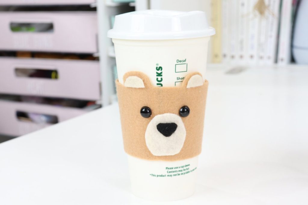 Image contains a tan felt bear coffee cozy on a white cup sitting on a white table.