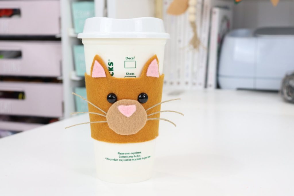 Image contains a coffee cozy made from felt in the shape of a cat’s face. The cozy is on a white to-go cup sitting on a white table.
