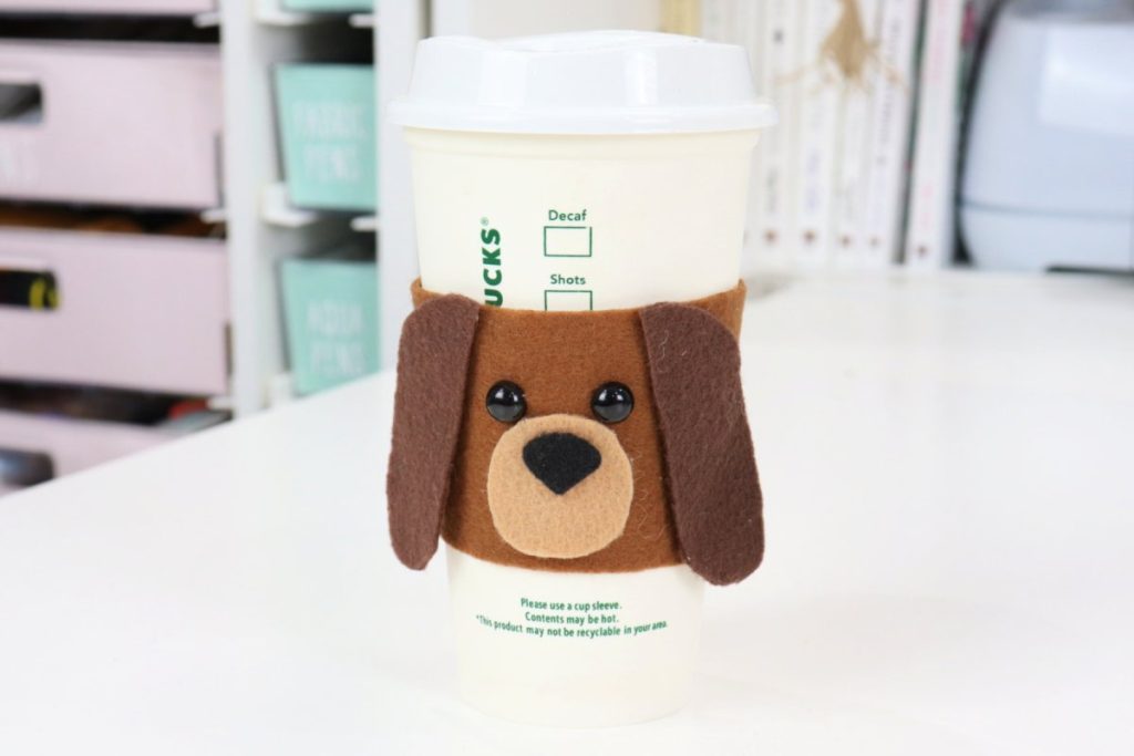 Image contains a coffee cozy made from brown felt to look like a dog’s face. The cozy is on a white to-go cup, sitting on a white table.