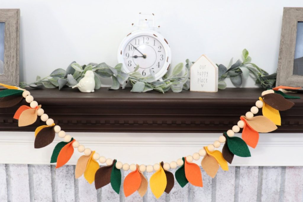 Image contains a garland made from natural wooden beads and felt leaves in shades of orange, brown, green, and yellow. It hangs on a whitewashed fireplace with a dark brown mantel.