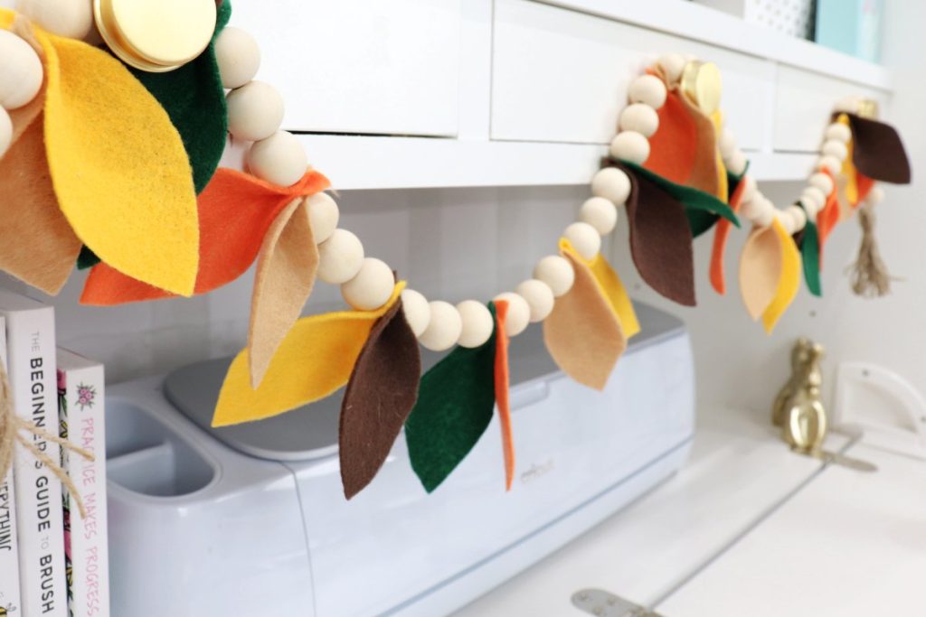 Image contains a garland made of natural wooden beads and felt leaves in shades of yellow, brown, green, and orange. It hangs on the drawers of Amy’s DreamBox organizer.