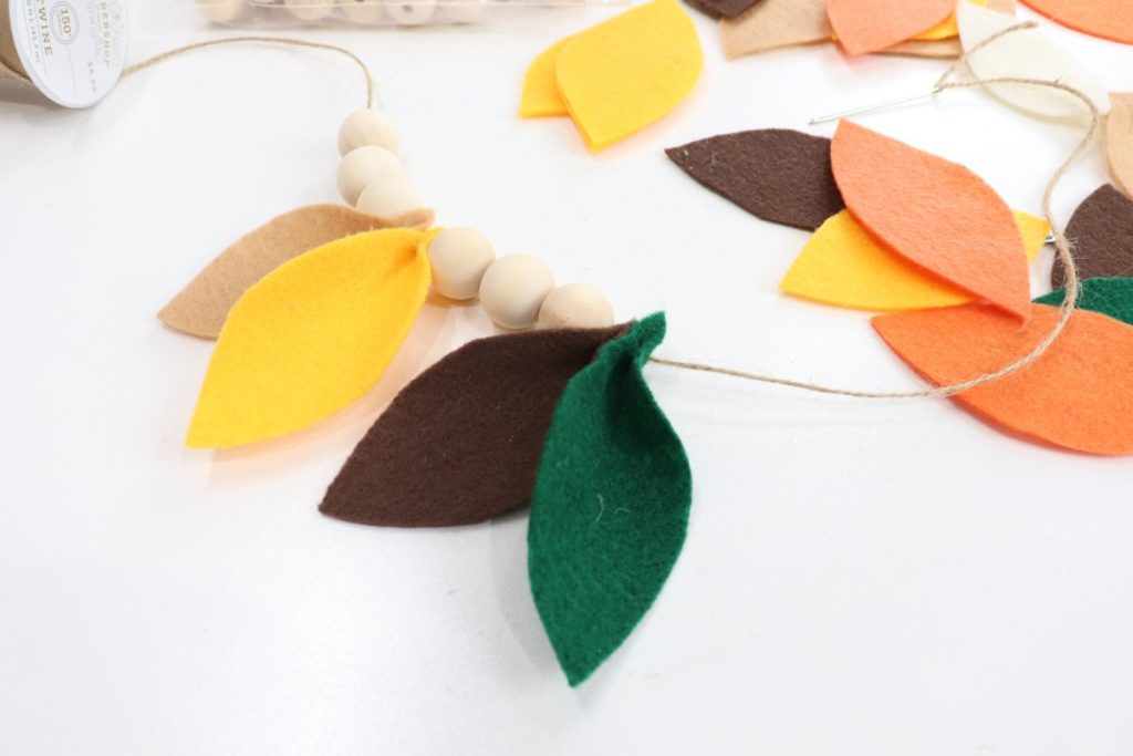 Image contains a piece of twine with three wooden beads followed by two felt leaves, three more beads, and two more leaves. Additional felt leaves in fall colors sit to the side on a white table.