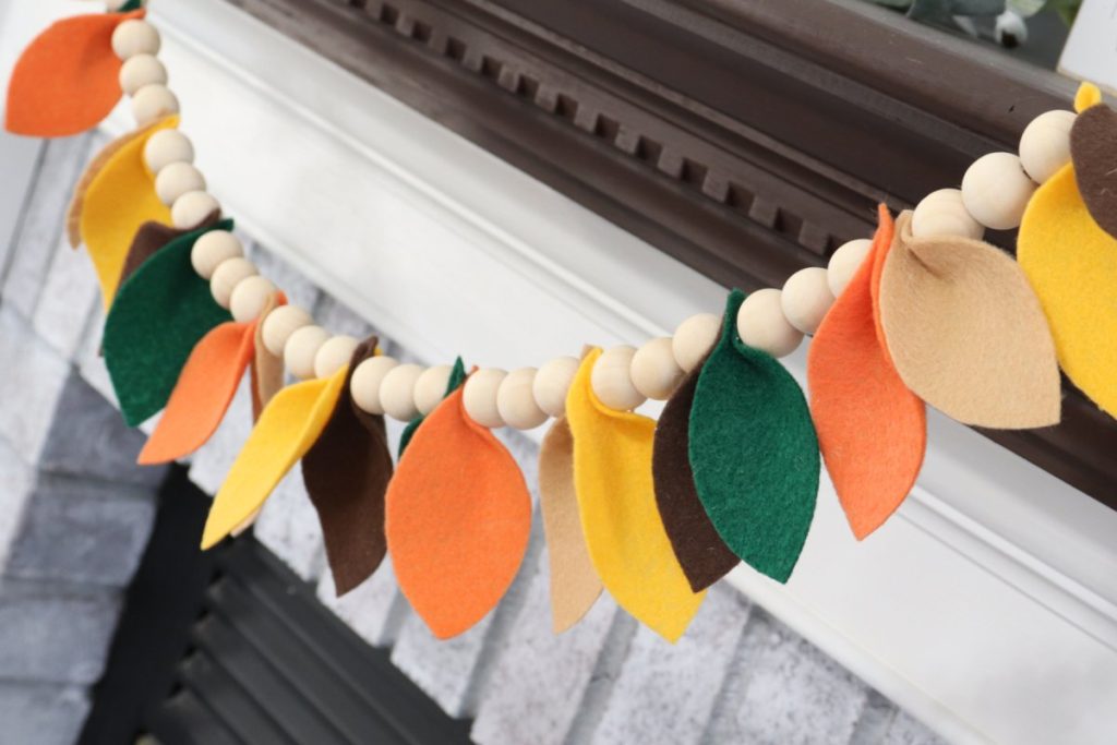 Image contains a fall garland made of wooden beads and felt leaves, hanging from a whitewashed fireplace.