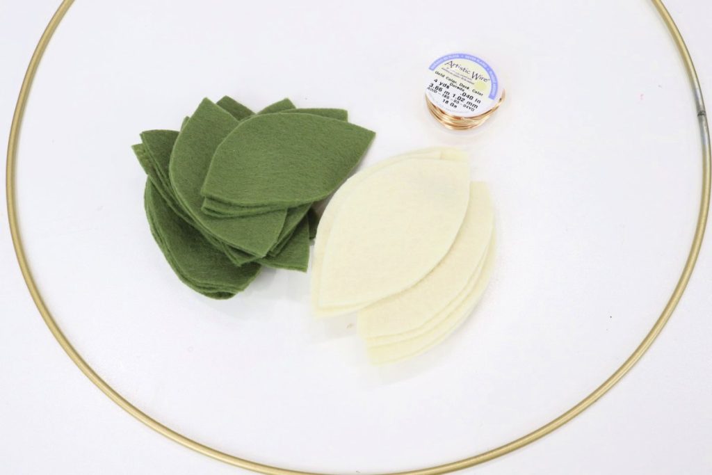Image contains a pile of green felt leaves and a pile of cream felt leaves inside a gold metal hoop. A spool of gold wire sits nearby.