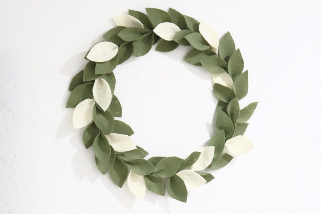 Image contains a wreath made of green and cream felt leaves on a white wall.