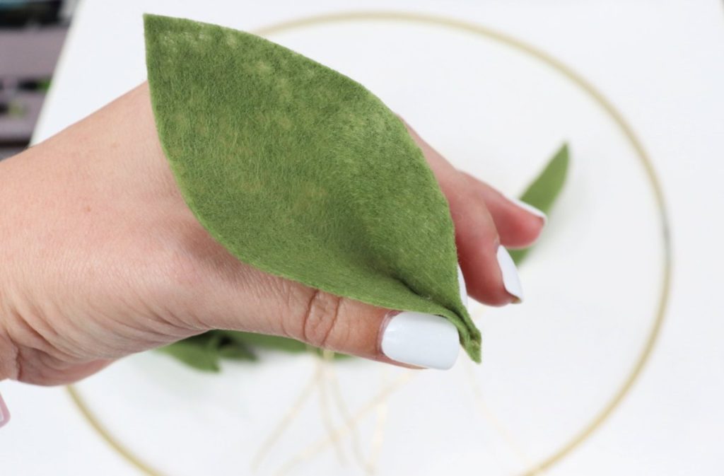 Image contains Amy’s hand pinching the bottom of a green felt leaf together. In the background, a metal hoop sits on a white table.