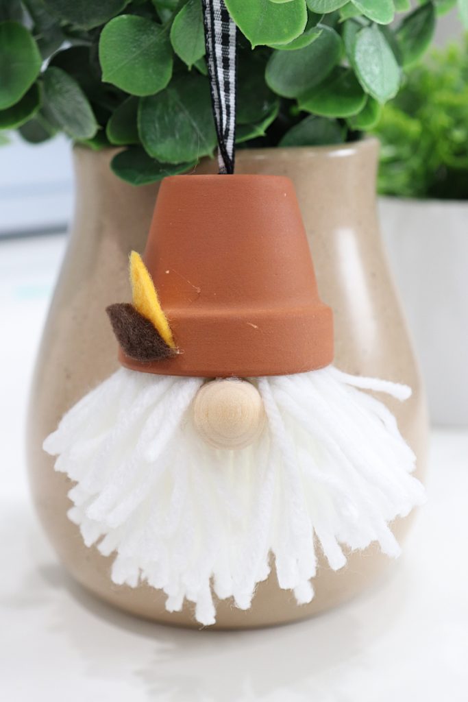 Image contains a gnome made from yarn, a wooden bead, a terracotta pot, and felt. It hangs in front of a faux plant in a brown vase.